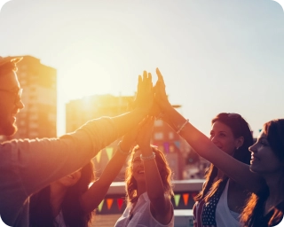 A group of people putting their hands together looking friendly with sunshine behind buildings in the background