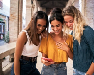 Three female coworkers looking at a persons phone with a sunny city background