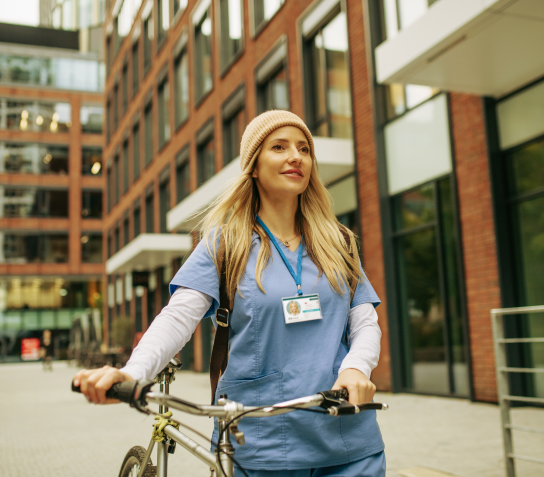 A female travel nurse in the city with a bike, knit cap, and blue scrubs