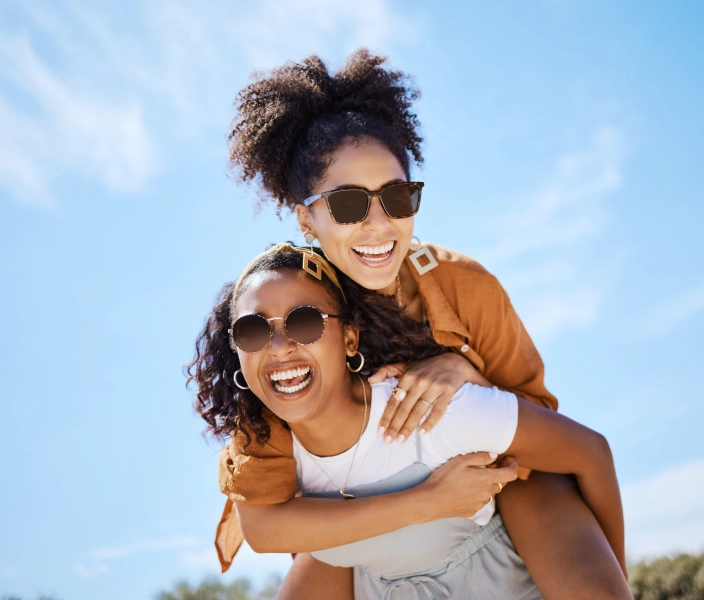 Two woman on a sunny day in sunglasses with one giving the other a piggyback ride