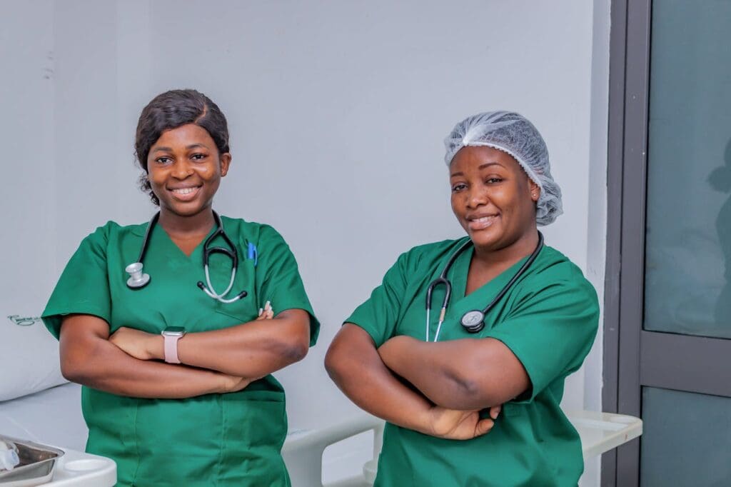 Two nurses wearing green scrubs, with their arms crossed smiling at the camera
