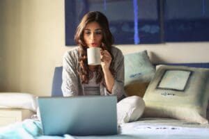 woman sitting in bed drinking from a mug while looking at a laptop